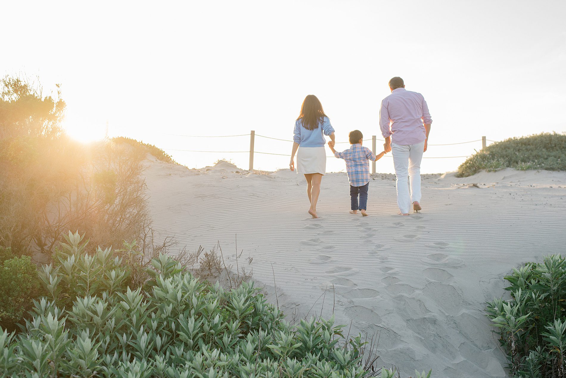 family of three hold hands walking up sand dune during Coastal Family Portraits in Carlsbad California 